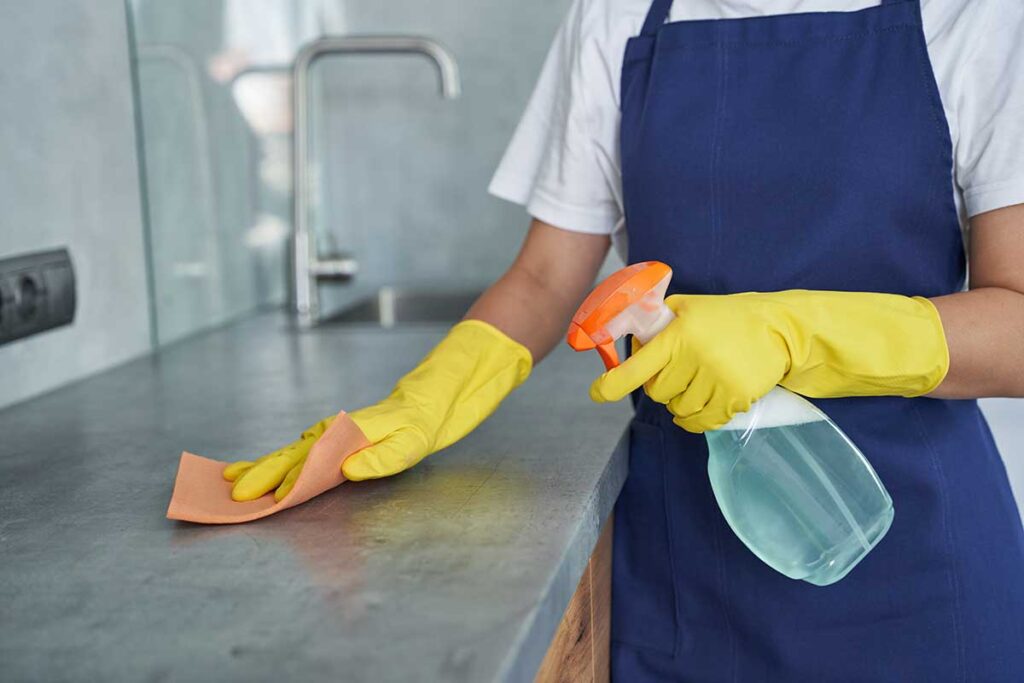 Person vacuuming a living room while cleaning supplies sit near a bright kitchen.