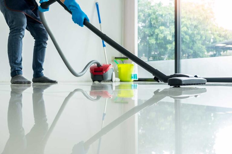 Person dusting shelves while a vacuum and cleaning cloths sit nearby in a bright home.