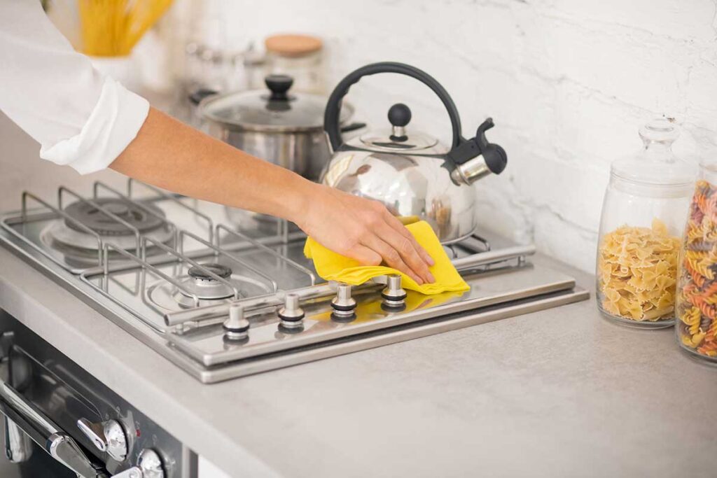 Person dusting a living room shelf while cleaning supplies and a vacuum wait nearby.